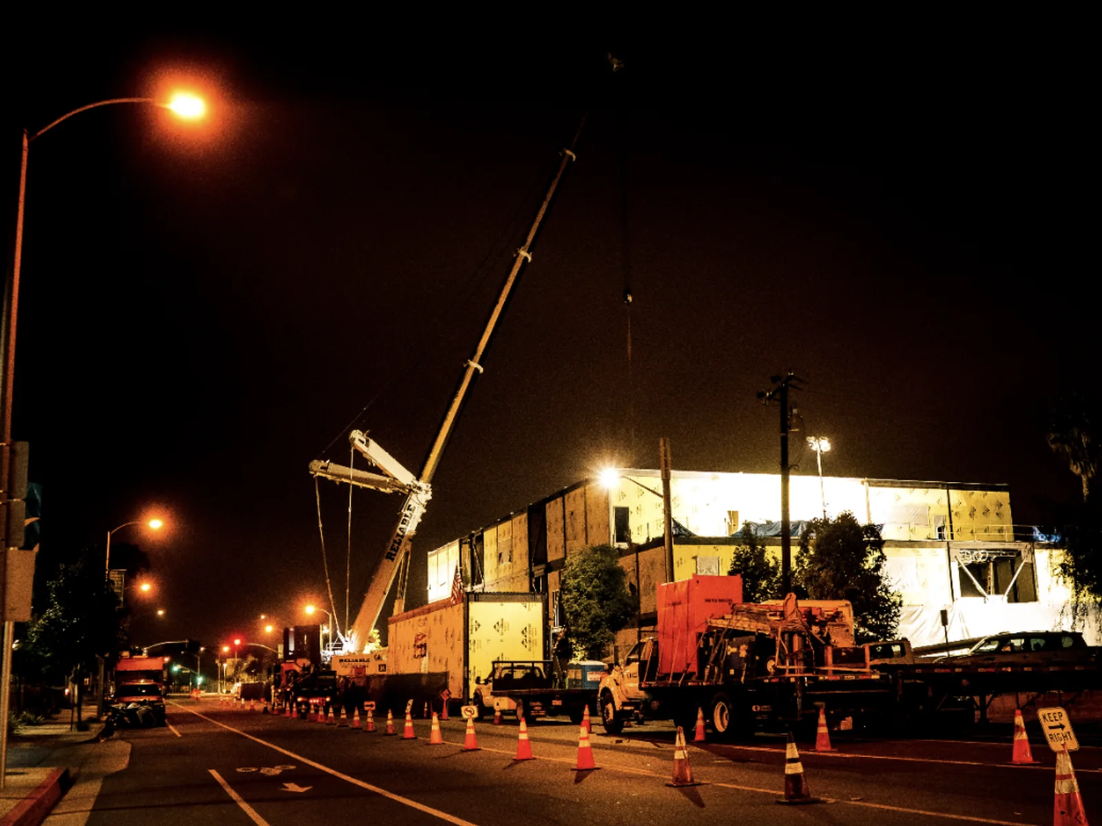 Nighttime crane operation setting modular building sections beside a city street